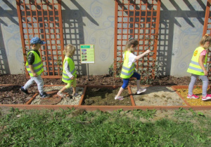 dzieci w seledynowych kamizelkach idą po ścieżce sensorycznej w ogrodzie obok drewnianych kratek, na których wija się pędy roślin pnących